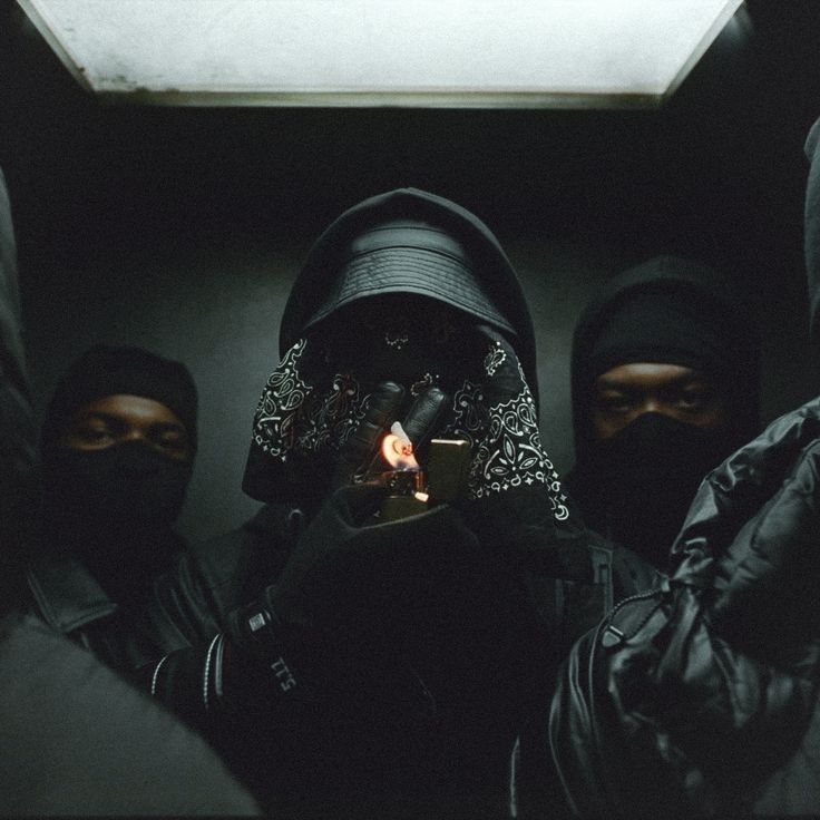 three people in black hoods and one is holding a lit candle while the other looks on