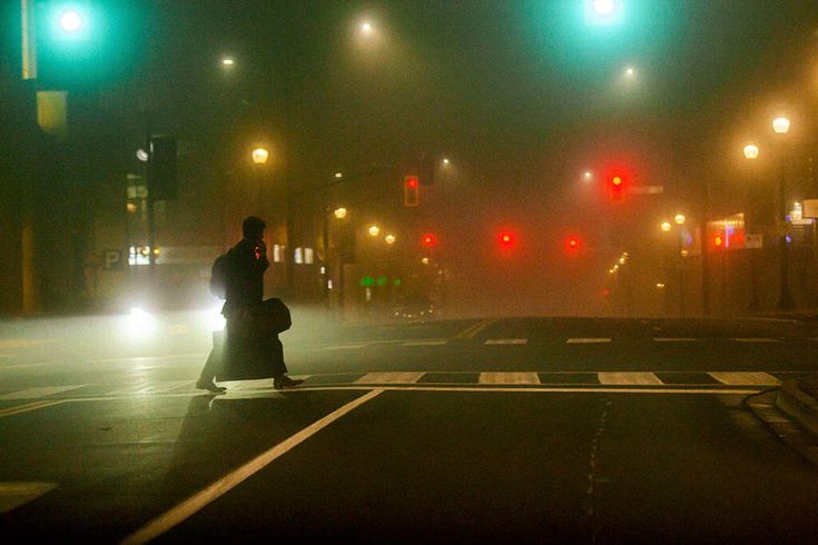 a person walking across a street at night with traffic lights in the background and fog on the ground