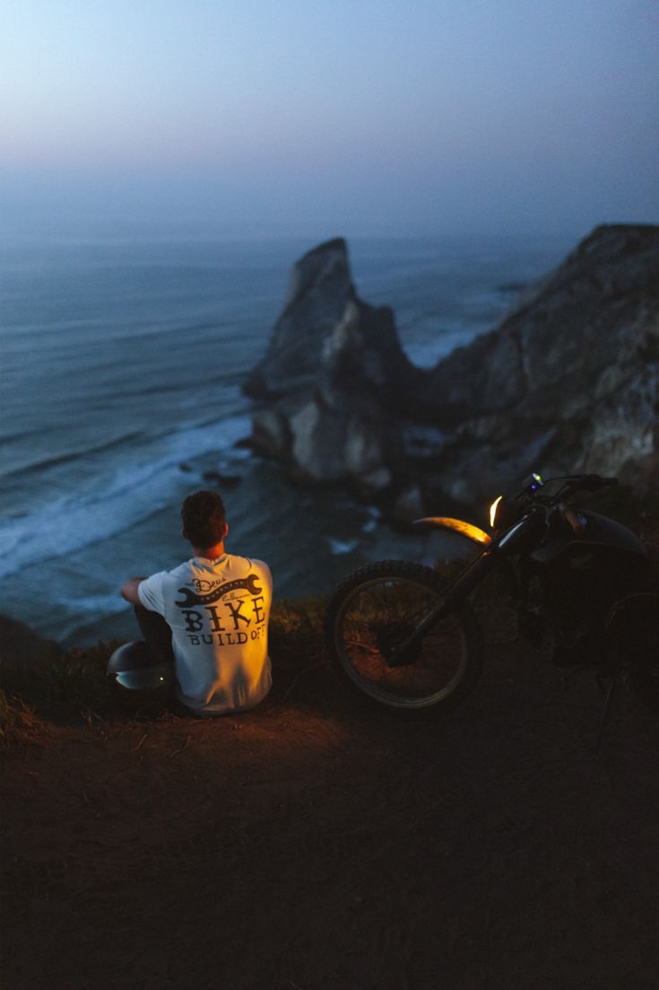 a man sitting on the side of a cliff next to a motorcycle near the ocean