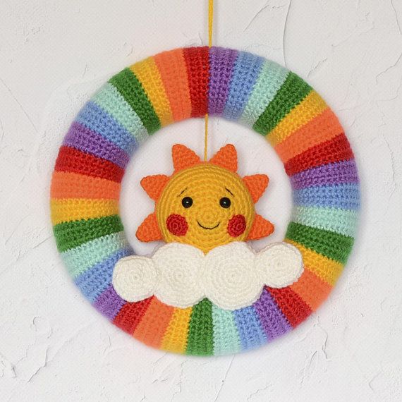 a crocheted wreath with a smiling sun and clouds hanging from the center, in front of a person's hand