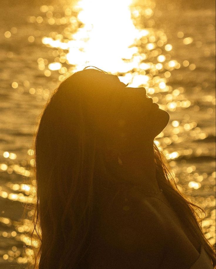 a woman standing in front of the ocean at sunset with her head tilted to the side