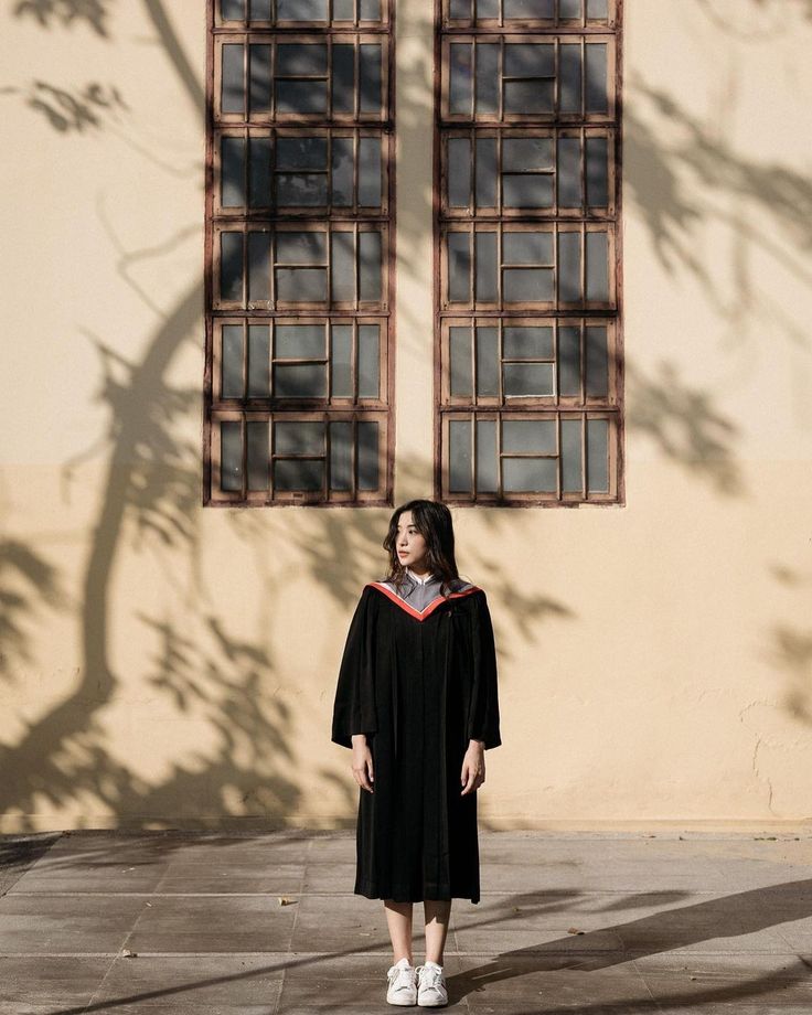 a woman wearing a graduation gown standing in front of a building with two large windows