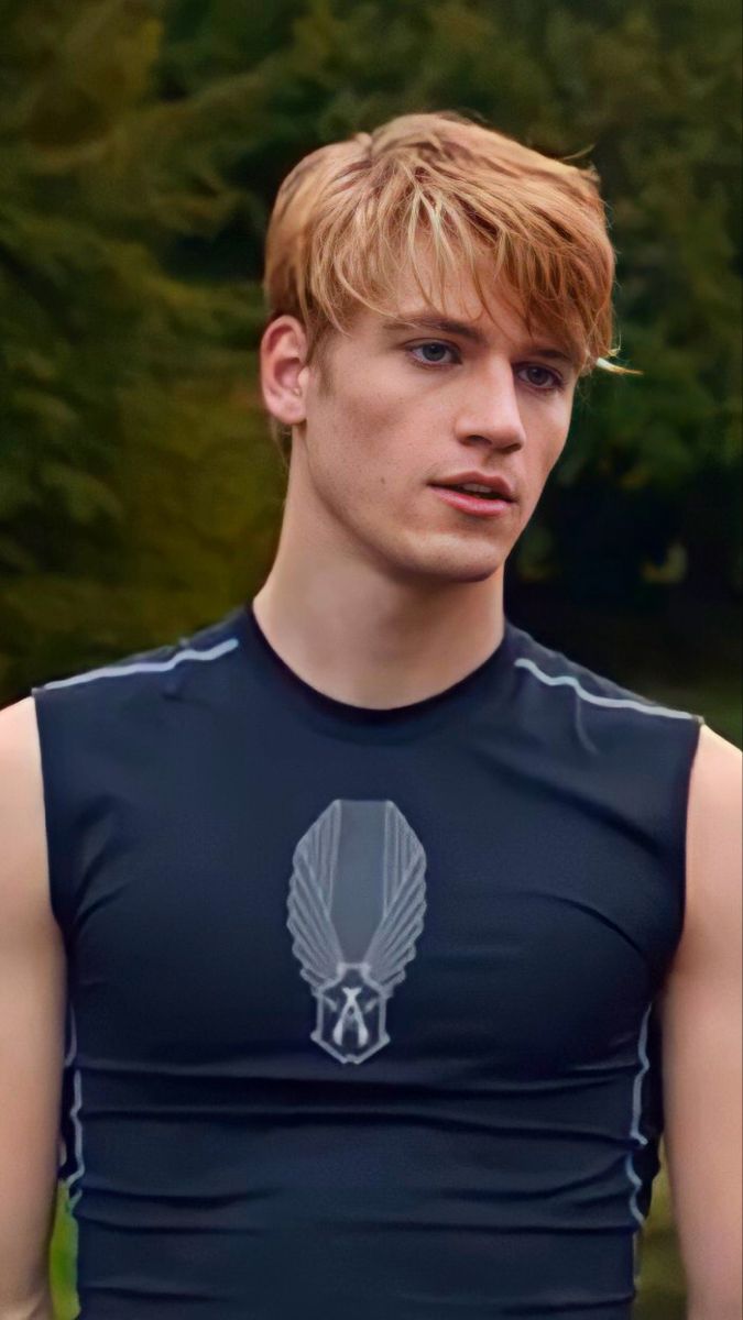 a young man with blonde hair wearing a black shirt and standing in front of trees