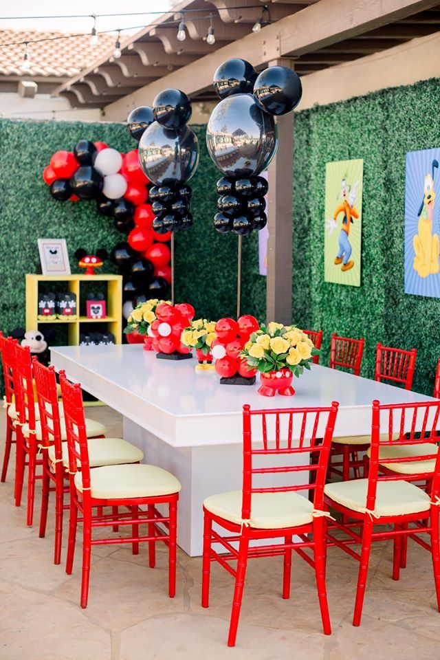 a table with red chairs and mickey mouse balloons hanging from it's ceiling in front of a green wall