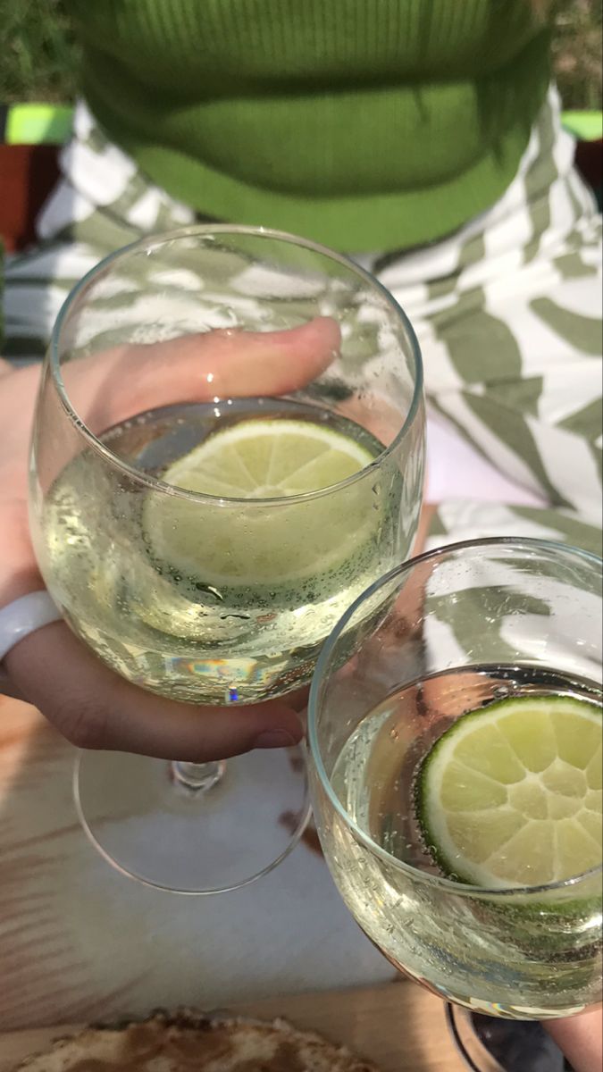 two people holding wine glasses filled with white wine and limes, while one person holds a slice of lemon in their hand