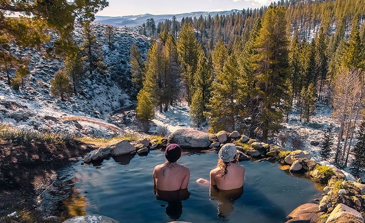 two people sitting in the middle of a pool surrounded by rocks and pine trees, with snow on the ground