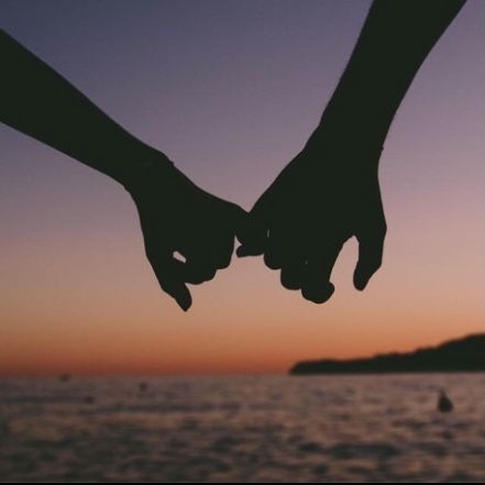 two people holding hands while standing in front of the ocean at sunset with boats in the background