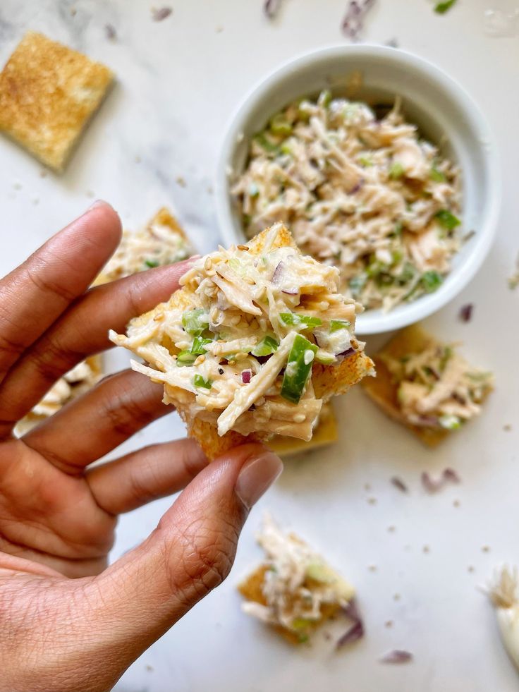 a hand holding a cracker with some food on it and a bowl of dip in the background