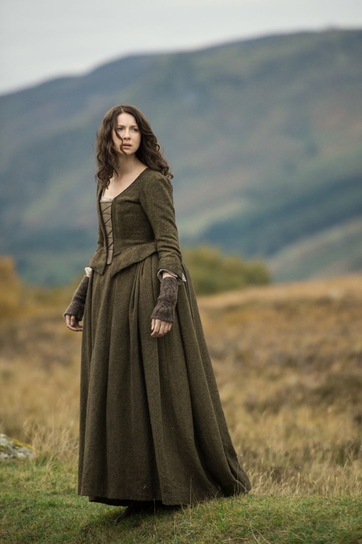 a woman in a long green dress standing on top of a grass covered field with mountains in the background