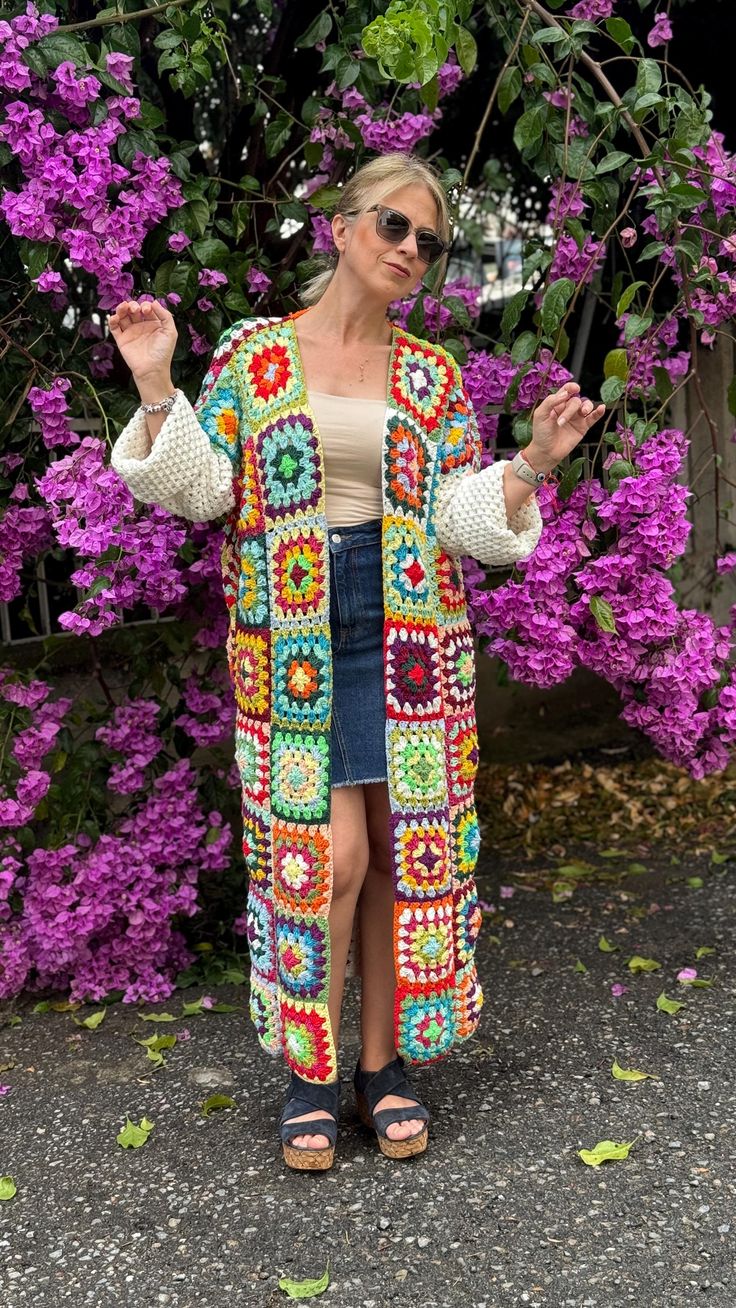 a woman standing in front of purple flowers wearing a colorful crochet cardigan