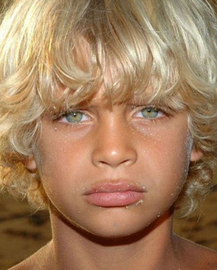 a young boy with blonde hair and blue eyes looks at the camera while he is covered in sand