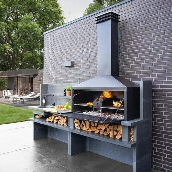 an outdoor bbq grill with wood stacked on the outside and brick wall behind it