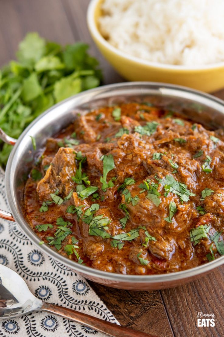 a metal bowl filled with food next to rice and cilantro leaves on a wooden table