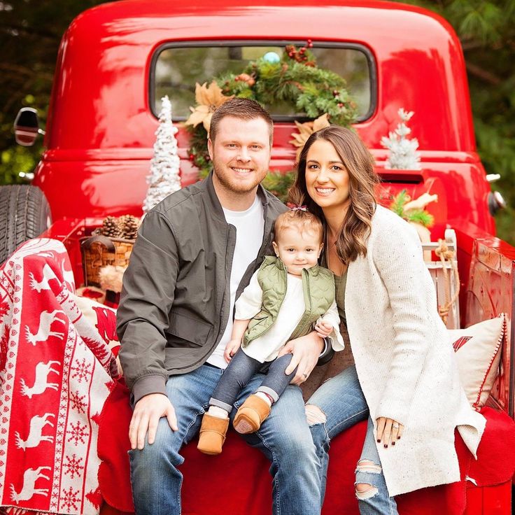 a man and woman sitting on the back of a red truck with a baby in their lap