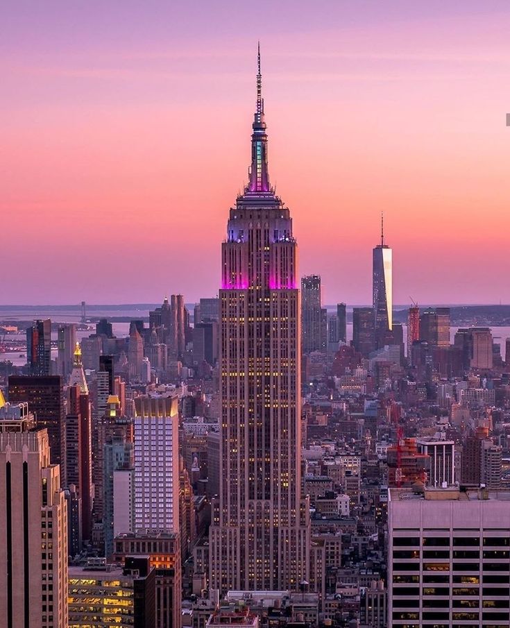 the empire state building lit up in purple and pink at sunset, as seen from top of the rock