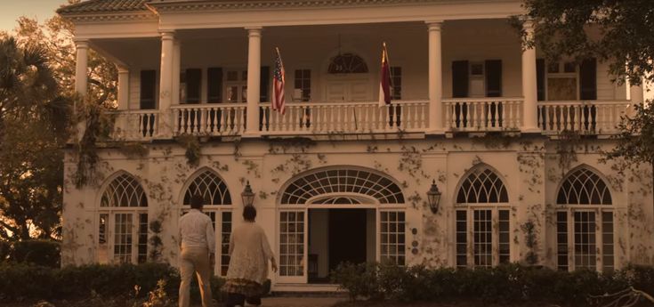 two people are walking in front of a large white house with columns and balconies