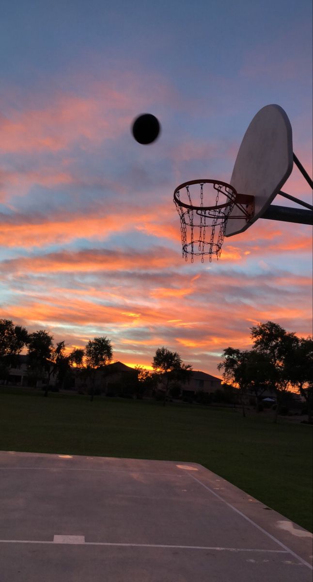 a basketball hoop with the sun setting behind it and a basketball flying through the air