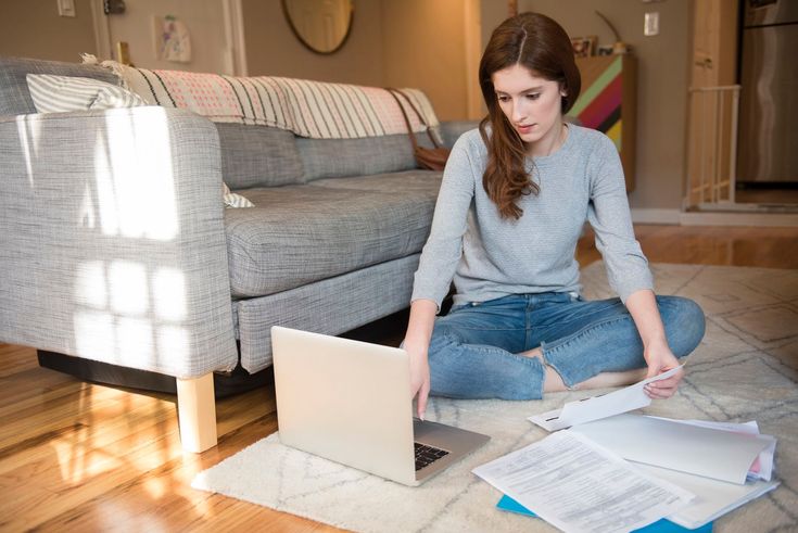 a young woman sitting on the floor working on her laptop computer while looking at paperwork