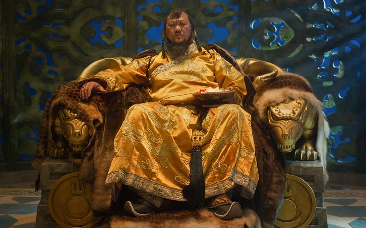 a man sitting on top of a golden chair next to two lion heads in front of a blue wall