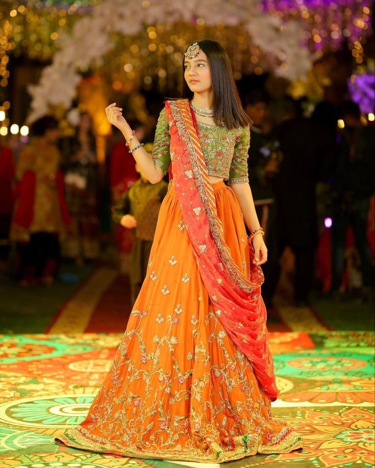 a woman in an orange and red lehenga standing on a stage with lights behind her