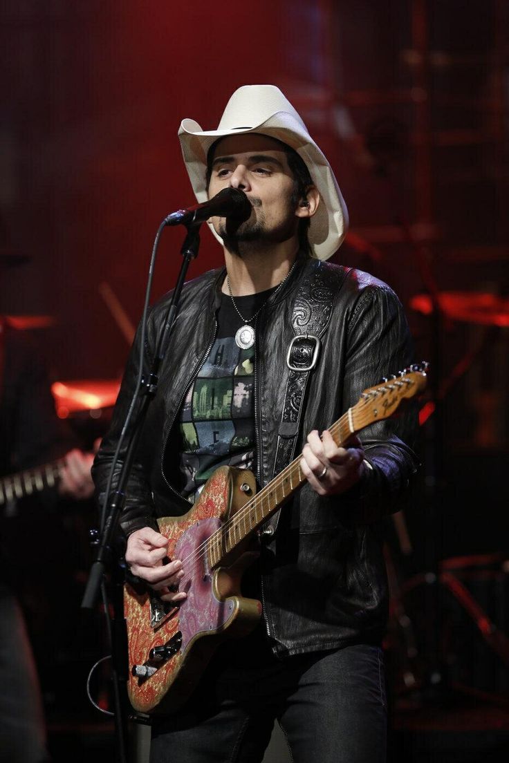 a man wearing a cowboy hat and holding a guitar in front of a microphone on stage