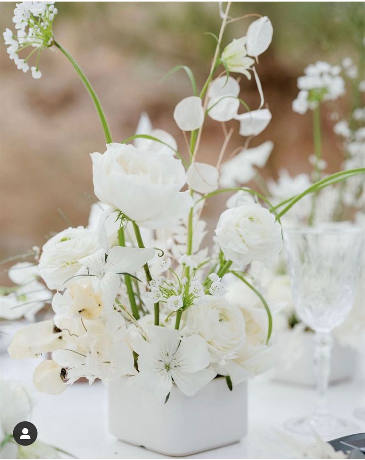 white flowers and greenery in a square vase on a table with wineglasses