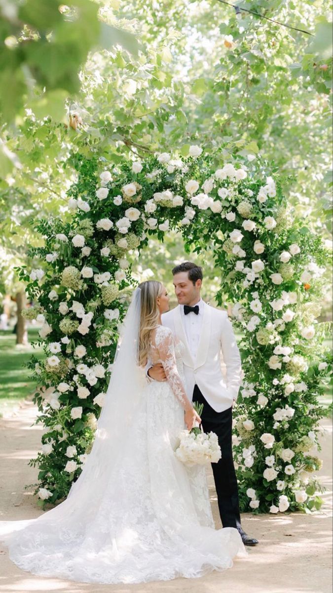 the bride and groom are standing in front of an arch with white flowers on it