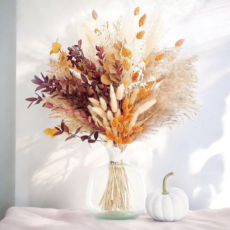 a vase filled with dried flowers on top of a white table next to a pumpkin