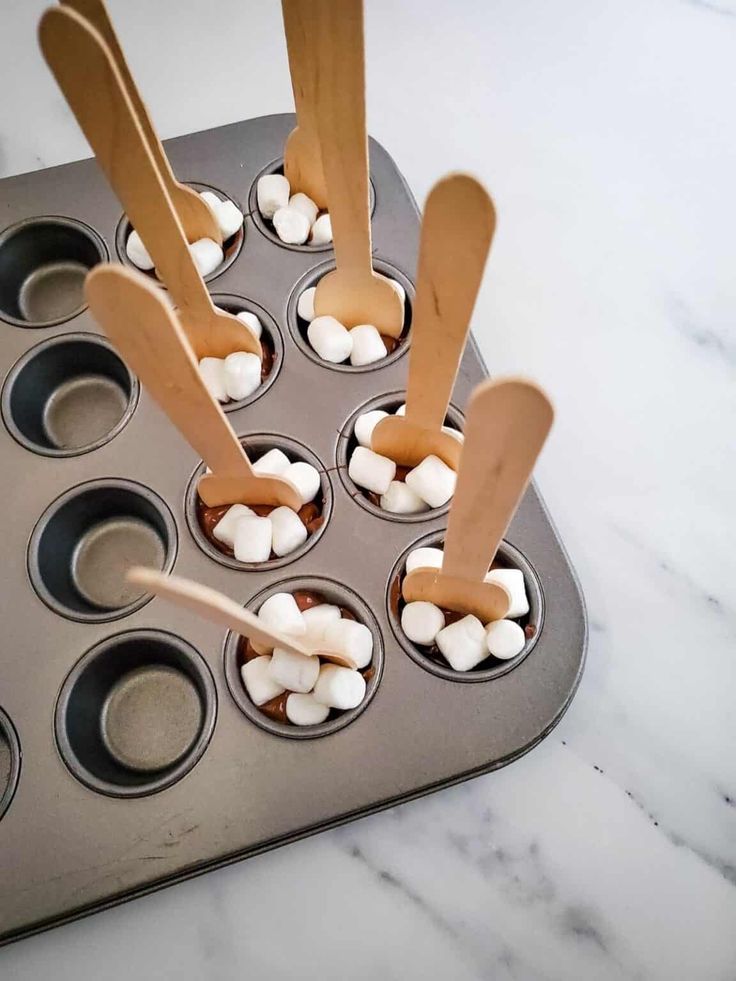 cupcake tins filled with marshmallows and wooden spoons on a marble countertop