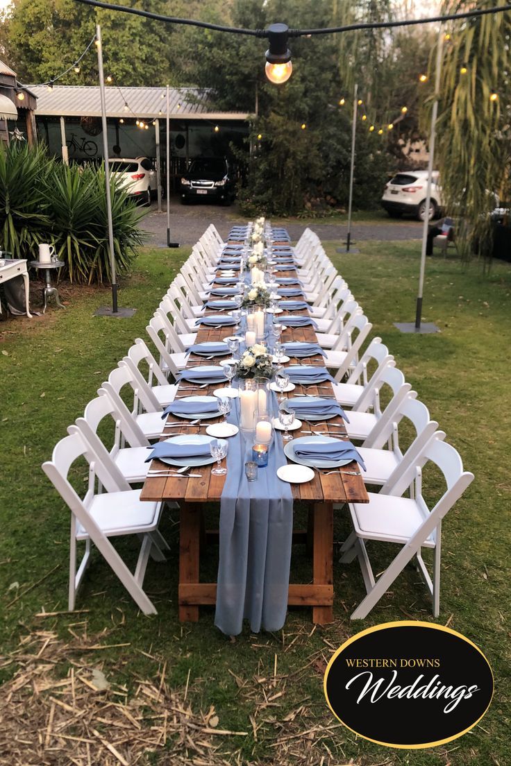 a long table set up with white chairs and blue linens for an outdoor wedding