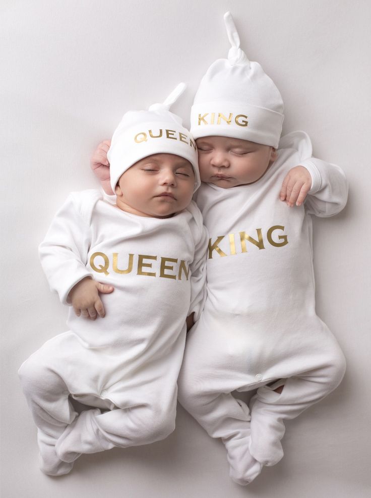 two baby babies wearing matching white outfits with gold lettering on them, one is sleeping while the other sleeps