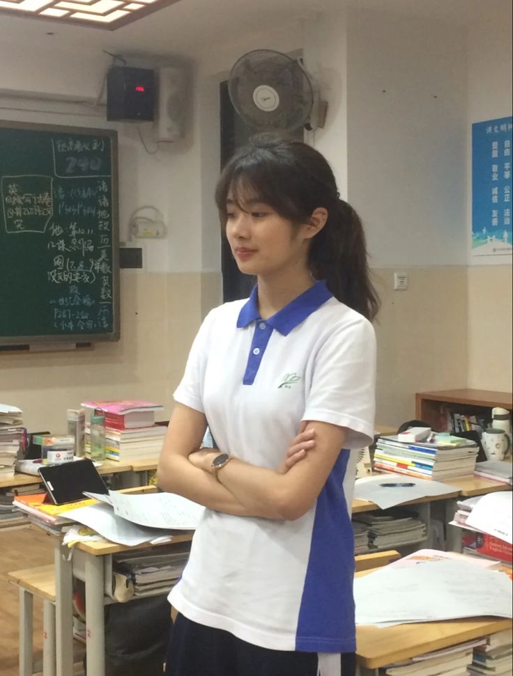 a woman standing in front of desks with books on it and a chalkboard behind her