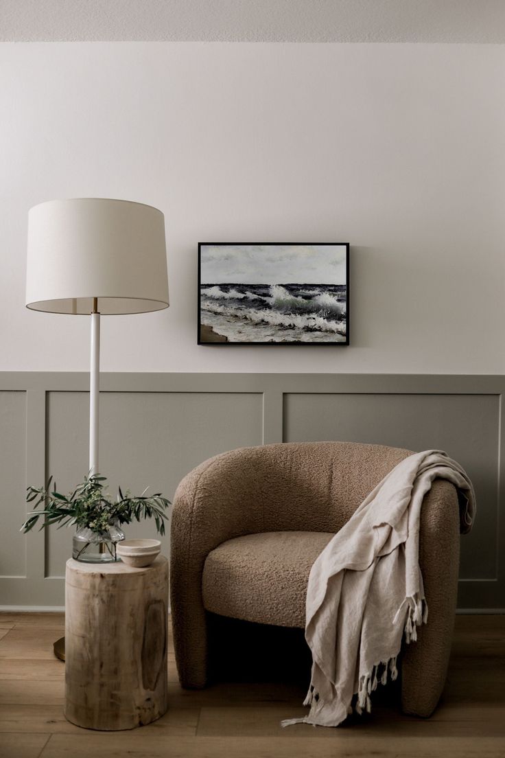 a chair with a blanket on it next to a lamp and a painting hanging on the wall