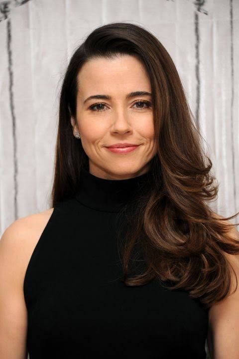 a woman with long brown hair wearing a black dress and smiling at the camera while standing in front of a white wall