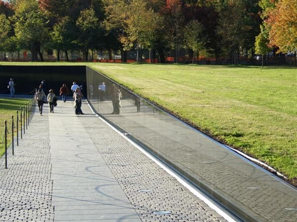 people are walking along the walkway in front of a reflecting pool and grassy area with trees