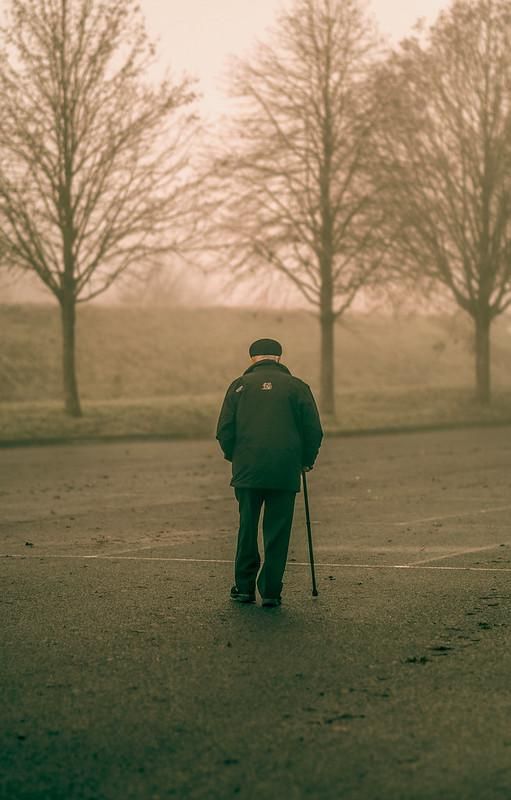 a man walking down the street with a cane in his hand and trees behind him