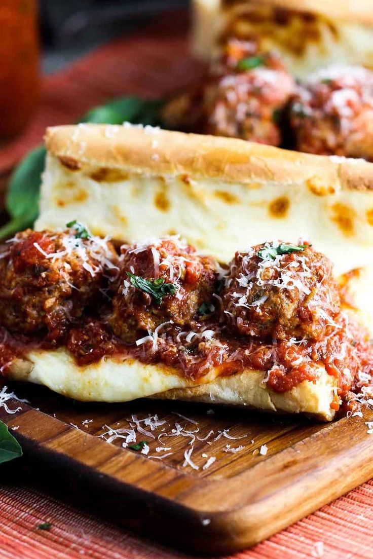 two meatball sub sandwiches on a wooden cutting board with basil leaves and sauce in the background