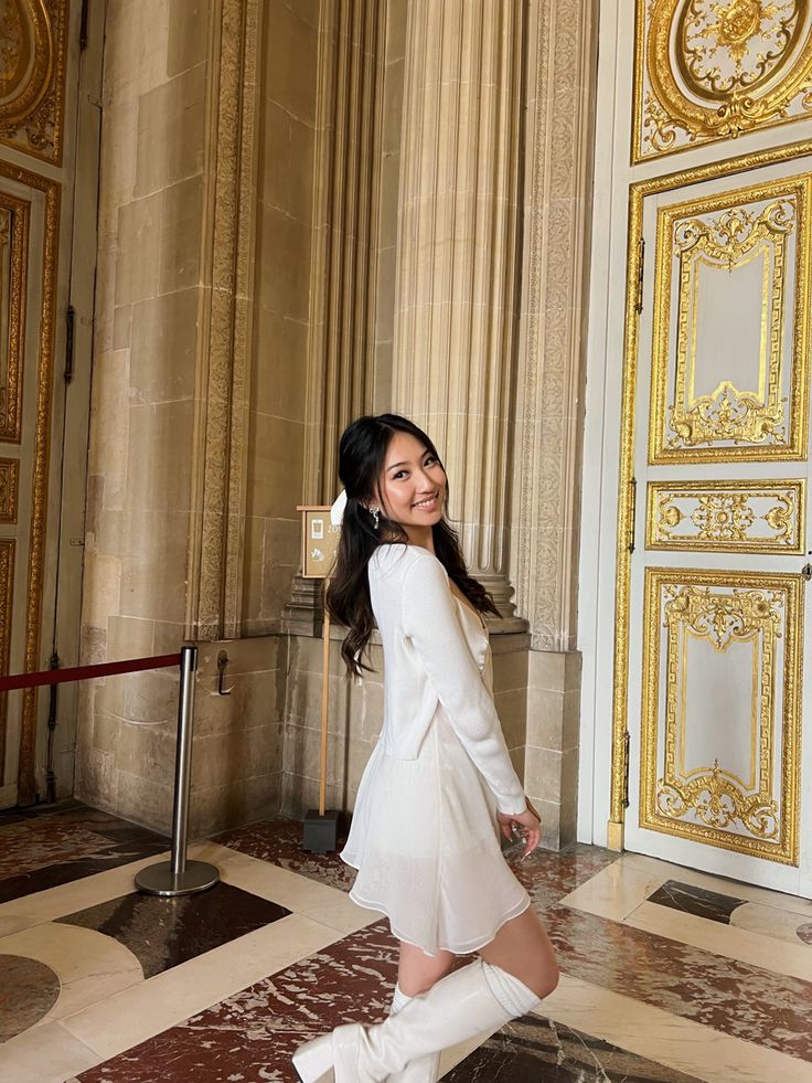 a woman in white dress and boots posing for the camera with gold doors behind her