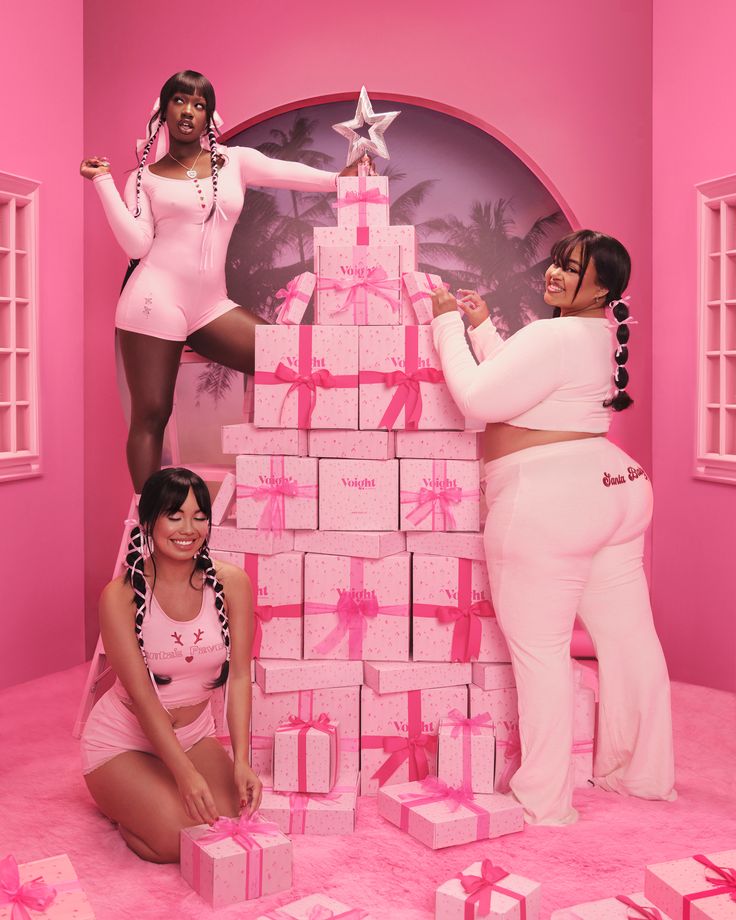 two women in pink outfits and one is leaning on a stack of gifts while the other woman stands next to her