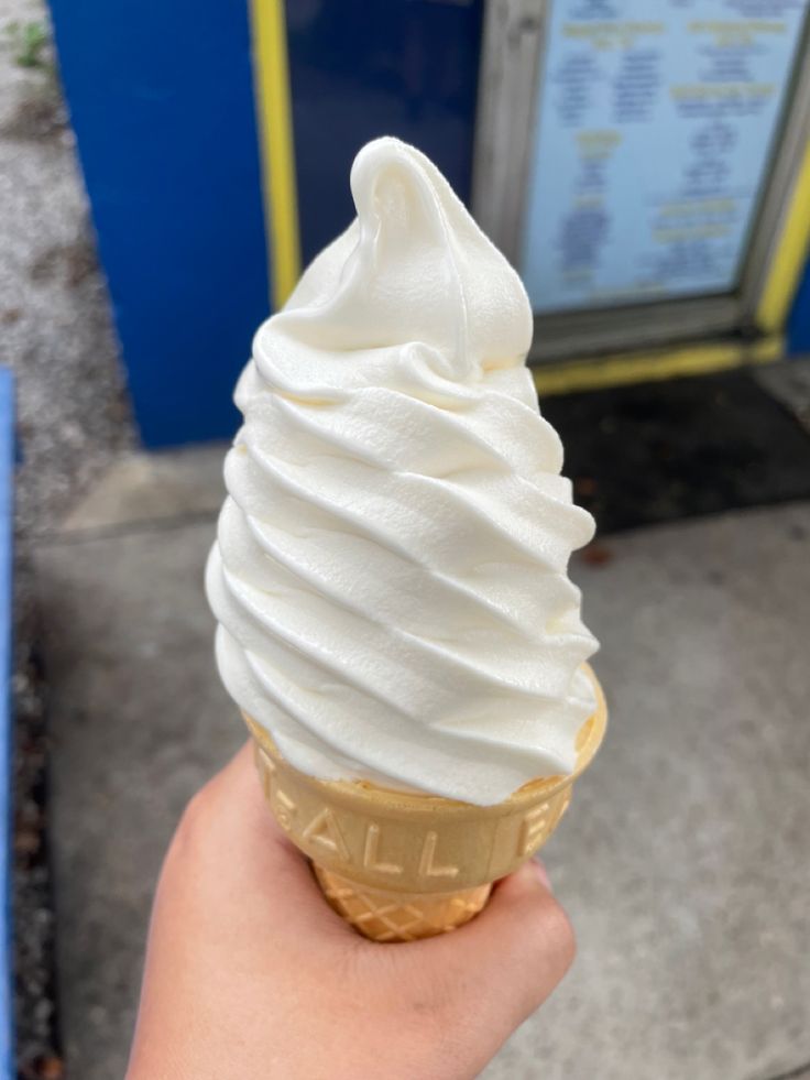 a hand holding an ice cream cone with whipped cream on top, in front of a sign