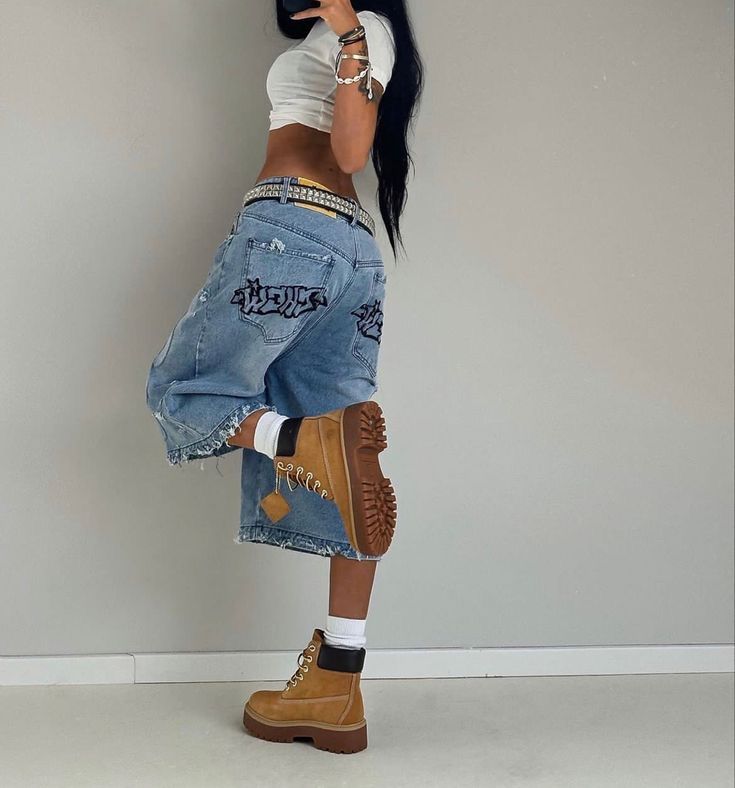 a woman with long black hair standing in front of a wall wearing ripped jeans and boots