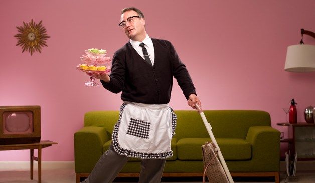 a man in an apron is holding a broom and flowers while standing next to a green couch