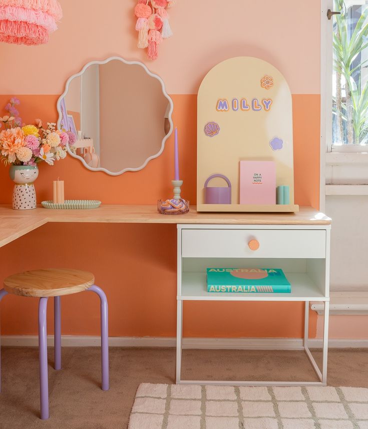 a desk with a mirror, stool and flower vase on it in a room that has peach walls