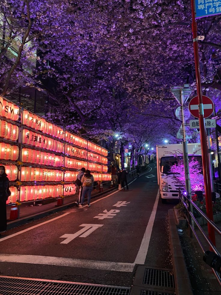 people are walking down the street in front of lighted signs and trees with purple lights on them