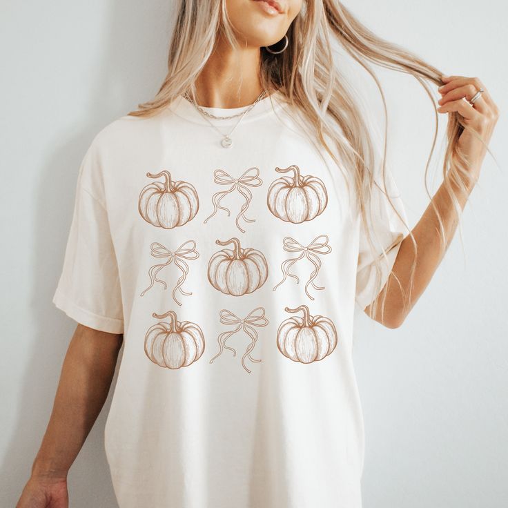 Discover Fall Charm with our Adorable Coquette Core Pumpkins and Bows T-shirt!  Embrace the essence of autumn with the perfect fall tee, a delightful addition to your seasonal wardrobe. Perfectly capturing the spirit of fall, this t-shirt features a charming design of whimsical pumpkins and bows, creating a playful yet elegant look. This shirt offers unparalleled comfort and softness, making it ideal for all-day wear. Its relaxed fit ensures a flattering silhouette that suits any body type. Whet Cute White Fall Shirt, Cute White Shirt For Fall, White Short Sleeve Shirt For Fall, Cute White Tops For Halloween, White Graphic Tee For Fall, White Tops For Fall, White Relaxed Fit Shirt For Fall, White Relaxed Fit T-shirt For Fall, Orange Graphic Tee For Fall