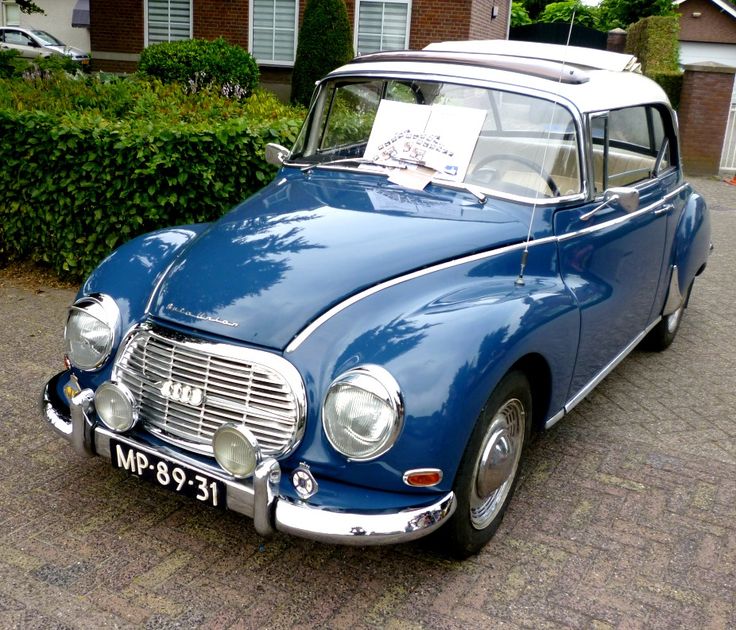 an old blue car is parked in front of some bushes and shrubs with a sign on the hood