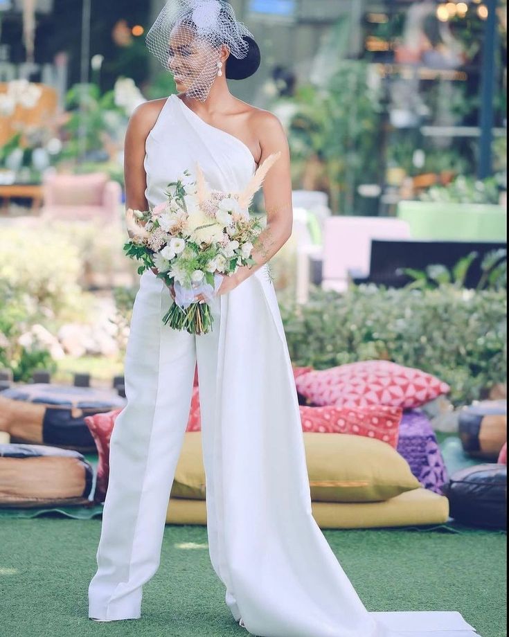 a woman in a white dress and veil standing next to a flower filled pillow on the ground