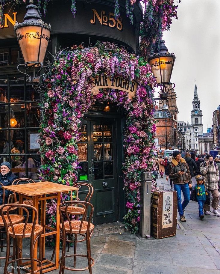 people are walking down the sidewalk in front of a building with flowers on it and tables outside