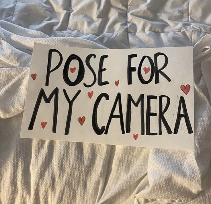 a sign that says pose for my camera on a bed with white sheets and pillows