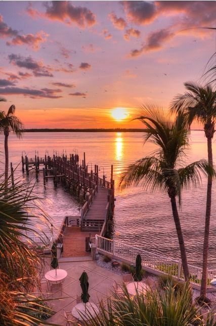 the sun is setting over the water and palm trees in front of a dock with umbrellas